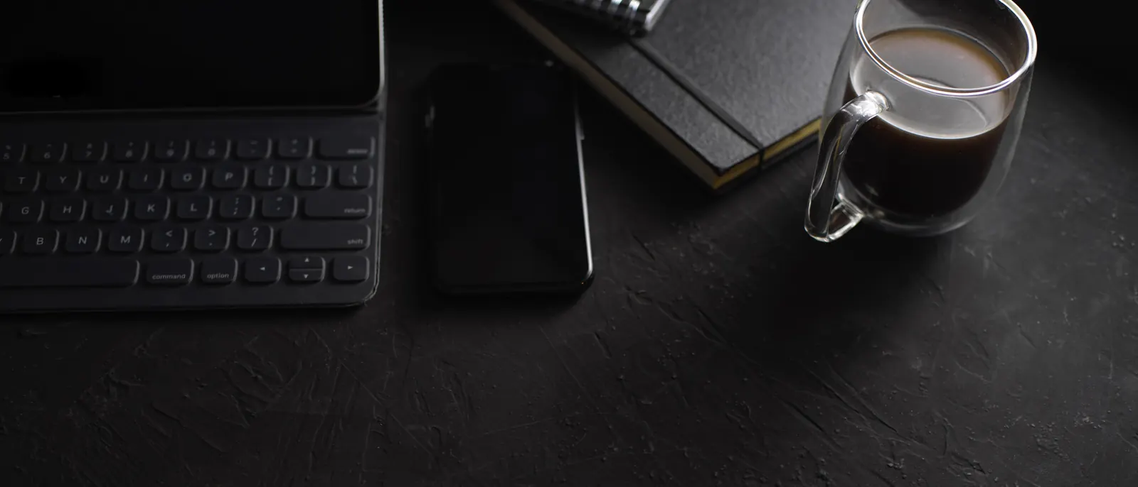 A dark desk with a keyboard, a notebook and a glass of coffee.