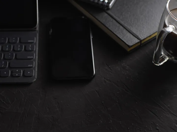 A dark desk with a keyboard, a notebook and a glass of coffee.