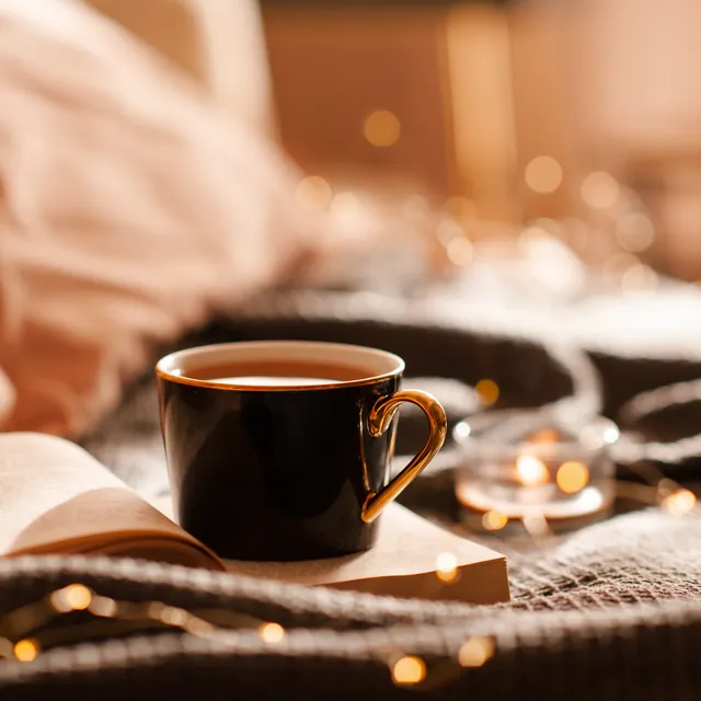 An open book and a dark mug on a bed, lit by a string of small lights.